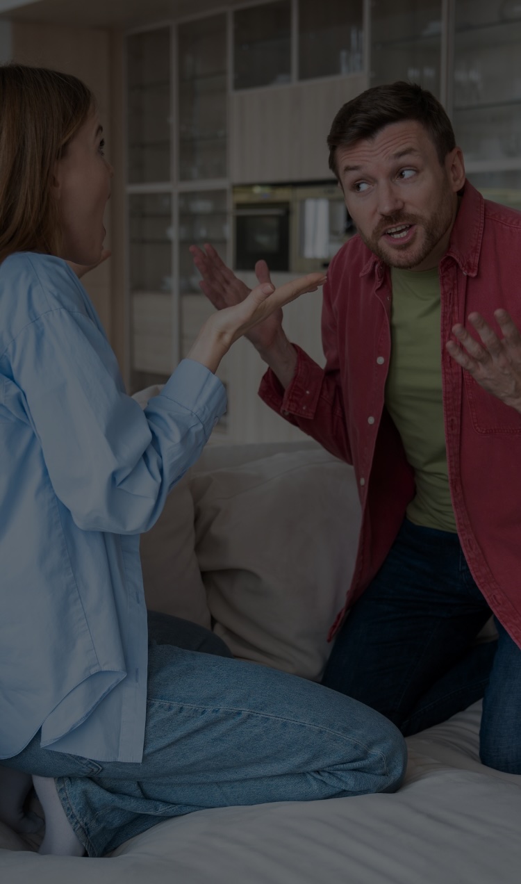 A couple argues on the couch with their hands in the air looking very animated.