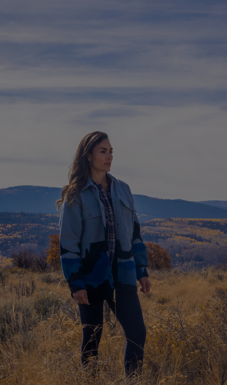 A woman stands in a field surrounded by the Colorado mountains.